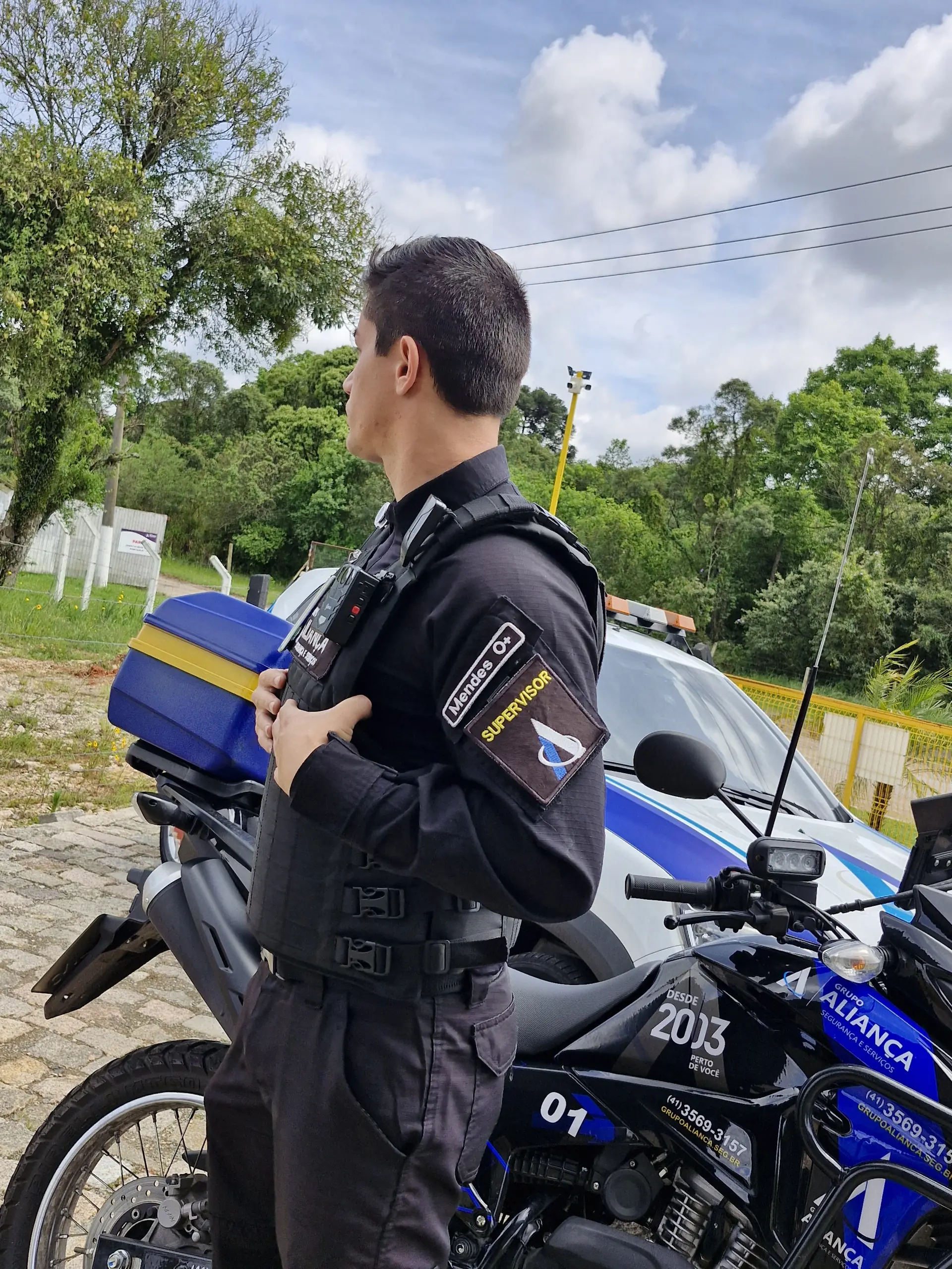 Agente tático do Grupo Aliança uniformizado, ao lado da moto e da viatura da empresa